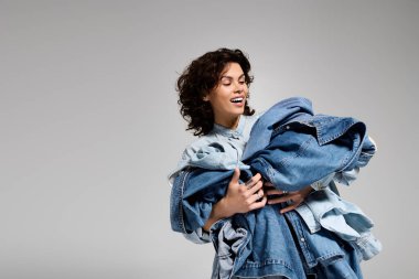 A joyful young woman with curly hair carries a trendy stack of denim clothes.