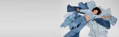 A young, stylish woman lies happily on a pile of various denim clothes in a vibrant studio setting.