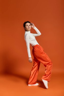 A stylish young woman poses elegantly against a warm orange backdrop.