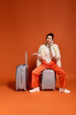 A trendy young woman in stylish orange pants and a chic outfit talks on her phone beside luggage.