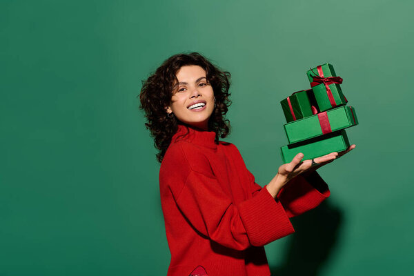 A woman in a red sweater smiles with wrapped gifts against a green backdrop.