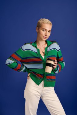 Moda kıyafetleri giymiş genç bir kadın kahve fincanı ile poz veriyor, modaya uygun bir görünüm sergiliyor..