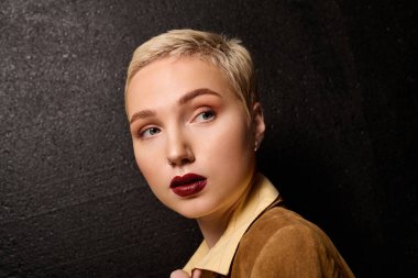 Kısa saçlı ve çarpıcı makyajlı zarif bir kadın, dokulu bir duvarın önünde zarifçe poz veriyor..