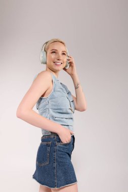 Genç bayan kulaklık takarken gülümsüyor, şık kot kıyafetini sergiliyor..