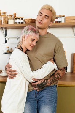 Bir çift genç aşık, parlak bir mutfakta kucaklaşır, neşe ve bağlantı yayar..