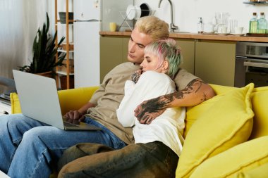 Genç bir çift, rahat oturma odalarında dizüstü bilgisayarlarıyla iş birliği yaparken birbirlerinin sıcaklığından zevk alır..