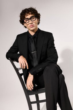 Şık giyinmiş şık bir genç adam, kendine güvenen bir sandalyede oturuyor. Çekicilik ve zarafet saçıyor..