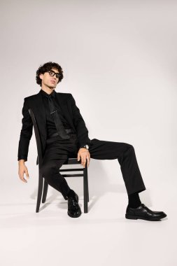 Gösterişli siyah takım elbiseli genç bir adam, sandalyede otururken çağdaş bir tarz sergiliyor..