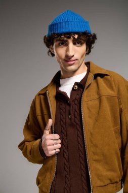 Modayı takip eden genç bir adam, kendine güvenen bir şekilde stüdyoda poz veriyor. Moda kıyafetleri ve mavi bereler giyiyor..