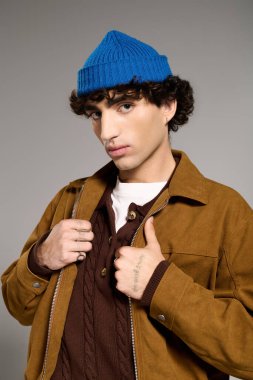 Rahat kahverengi ceketli ve mavi bereli yakışıklı bir adam çağdaş bir stüdyoda rahatça poz veriyor..