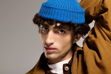 Genç bir adam şık kıyafetler ve canlı mavi bereyle stüdyo ışığı altında cazibe ve tarz yayar..