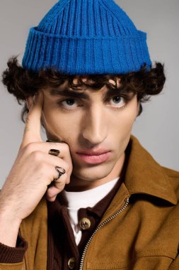 Kıvırcık saçlı genç adam mavi bir bere takıyor ve kendine has tarzını sergiliyor..