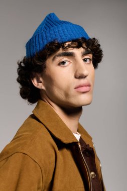 Yakışıklı genç bir adam kendine güvenerek duruyor, modaya uygun mavi bir bere ve şık bir kahverengi ceket giyiyor..