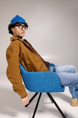 Genç ve yakışıklı bir adam şık kıyafetini sergiliyor. Canlı bir sandalyede rahatça oturuyor..