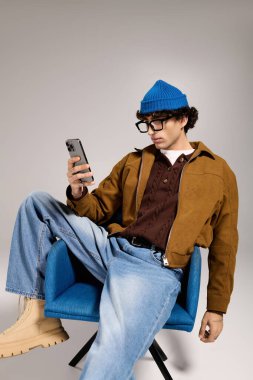 Zeki giyimli genç adam mavi bir sandalyede oturmuş, dikkatle akıllı telefonuna bakıyor..