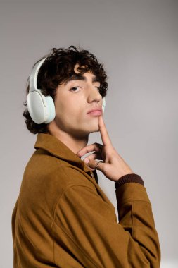 Kıvırcık saçlı genç bir adam şık giyinmiş ve kulaklık takmış düşünceli bir şekilde poz veriyor..