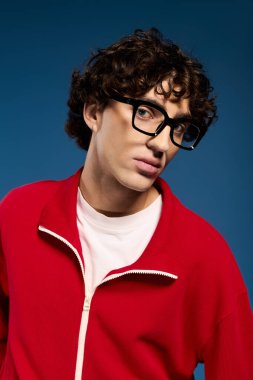 Kıvırcık saçlı genç bir adam modern moda kırmızı elbiseli ve kalın gözlükleri sergiliyor..