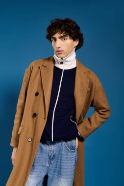 Moda bir ceket ve günlük giysiler içinde genç bir adam cesur mavi bir arka plana karşı güvenle poz verir..