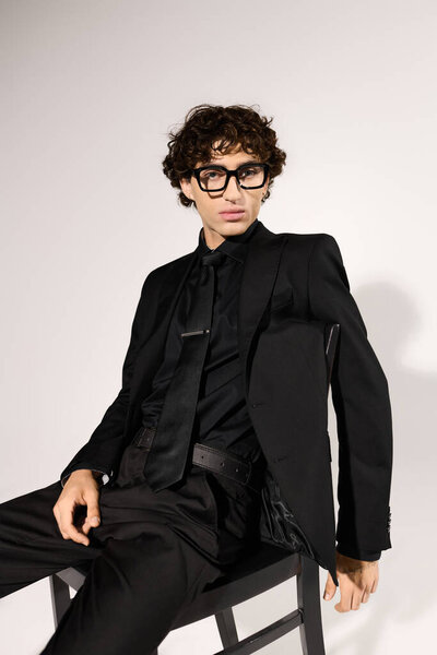 A young handsome man in sleek black attire poses confidently against a minimalist backdrop.