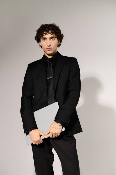 Молодой человек излучает шарм и изысканность, держа в руках ноутбук, одетый в гладкий черный наряд.
