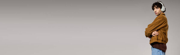 A young man stands confidently, dressed in fashionable attire and enjoying music through headphones.