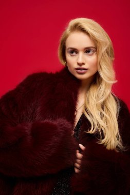 Genç sarışın bir kadın şık kırmızı kürkünü canlı bir arka planda sergiliyor..