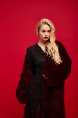 Kırmızı kürklü genç bir sarışın canlı kırmızı bir arka plana karşı güvenle poz veriyor..