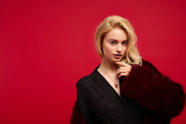 Красивая блондинка уверенно позирует в шикарном наряде на ярком фоне.