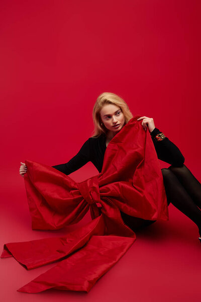 Молодая блондинка изящно позирует в черном вечернем наряде на праздничном красном фоне.