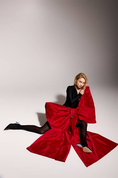 Молодая женщина в стильном черном наряде элегантно сидит на сером фоне.