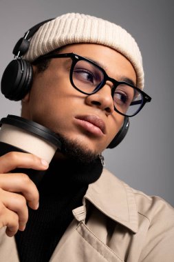 Moda kıyafetleri içinde şık bir genç, kahve yudumluyor ve modern bir ortamda kulaklık takıyor..