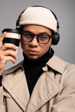 Kendine güvenen genç bir adam elinde kahve fincanı, spor kıyafetleri ve şık kulaklıklar tutuyor..