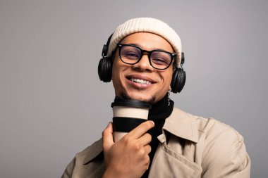 Çekici bir gülümsemesi olan genç adam modaya uygun kıyafetler ve kulaklık takıyor. Elinde kahve fincanı var..