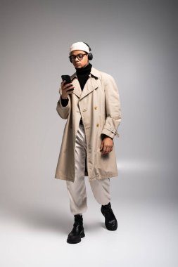 Moda kıyafetleri içindeki genç bir adam, şık bir ortamda akıllı telefonuna dalmış, kendinden emin bir şekilde duruyor..