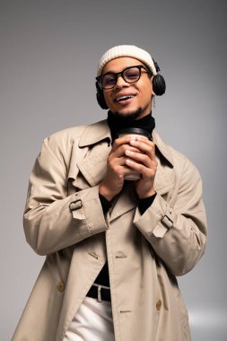 Genç adam, arka planda şık kıyafetler ve kulaklıklar takmış, sıcak bir içki tutuyor..