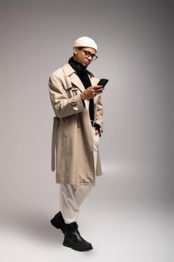 Yakışıklı genç bir adam kendine güvenerek duruyor, şık bir trençkot ve bereyle telefonunu kontrol ediyor..