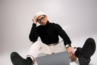 Siyah kazaklı ve beyaz pantolonlu şık bir genç adam laptopuyla düşünceli bir şekilde oturur..