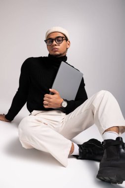 Moda kıyafetli yakışıklı genç adam sakince oturur, elinde bir dizüstü bilgisayarla çağdaş bir alanda..