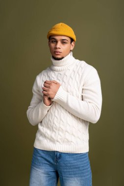 Genç adam rahat bir süveter ve temiz bir bereyle tarafsız bir zemine karşı güvenle duruyor.