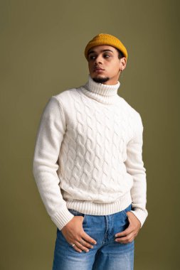 Şık bir süveter ve rahat bir kot giyen kendine güvenen genç bir adam yeşil bir fona karşı poz veriyor..