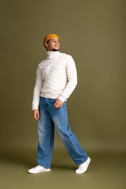 Rahat bir süveter ve rahat bir kot giyen genç adam sessiz bir arkaplanda rahatça poz veriyor..