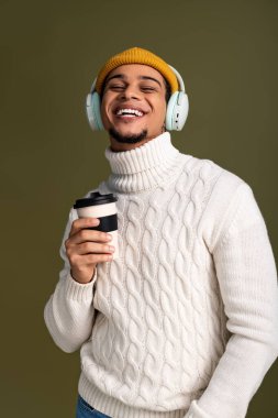 Rahat bir süveter giyen genç adam kahve fincanı ve kulaklıkla güvenle gülümsüyor..