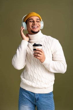 Tarz sahibi genç adam kahve fincanı tutarken gülümsüyor ve rahat bir kazak ve şapka giyiyor..