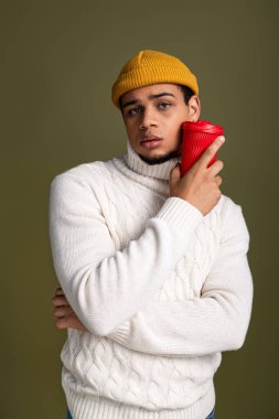 Genç bir adam sade bir arka planda rahat bir süveter ve renkli bir şapkayla tarzını sergiliyor..