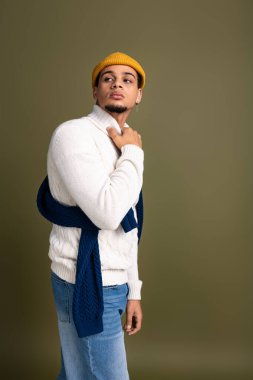 Yakışıklı genç adam rahat bir süveter ve şık bir şapkayla sessiz bir arkaplanda rahatça poz veriyor..