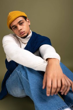 Yakışıklı genç adam, şık kıyafetler ve parlak bir bere giyerek öylece yerde oturuyor..