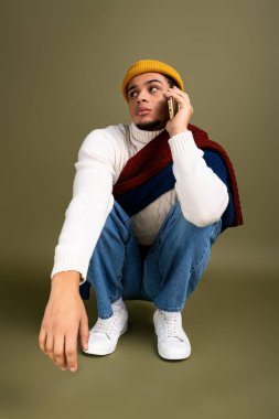 Şık bir genç adam yere çömeliyor, kendine güvenerek telefonda konuşuyor, renkli bir kıyafet giyiyor..