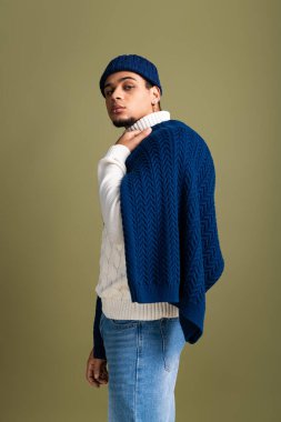 Çekici genç adam kendine güveni tam bir şekilde şık örgü elbisesiyle poz veriyor, yumuşak tonlarda modern moda yayıyor..