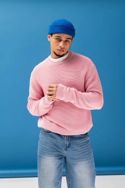 Pembe kazaklı ve mavi bereli şık bir genç adam parlak mavi bir fona karşı poz veriyor..
