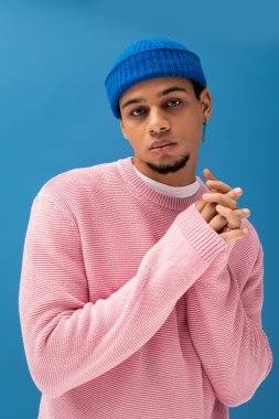 Şık bir genç adam pembe bir süveter ve mavi bir bere takıyor, elleri bağlı..